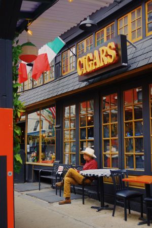 Person in cowboy hat relaxes outside a cigar shop storefront with illuminated sign and Italian flags hanging above.
