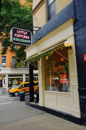 Little Cupcake Bakeshop storefront with vintage neon sign on a New York City street corner with yellow taxi visible.