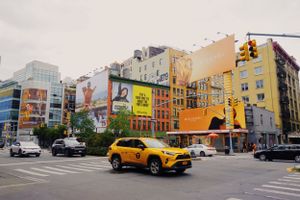 A yellow taxi drives past colorful buildings and murals on a busy street corner in New York City.