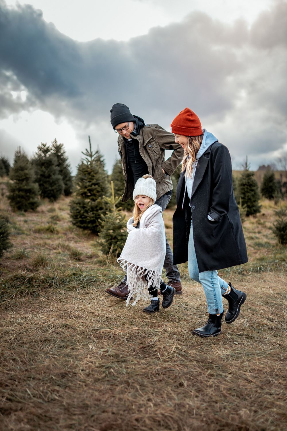 Three people walking together through an evergreen tree farm on a cloudy winter day wearing warm knit beanies.