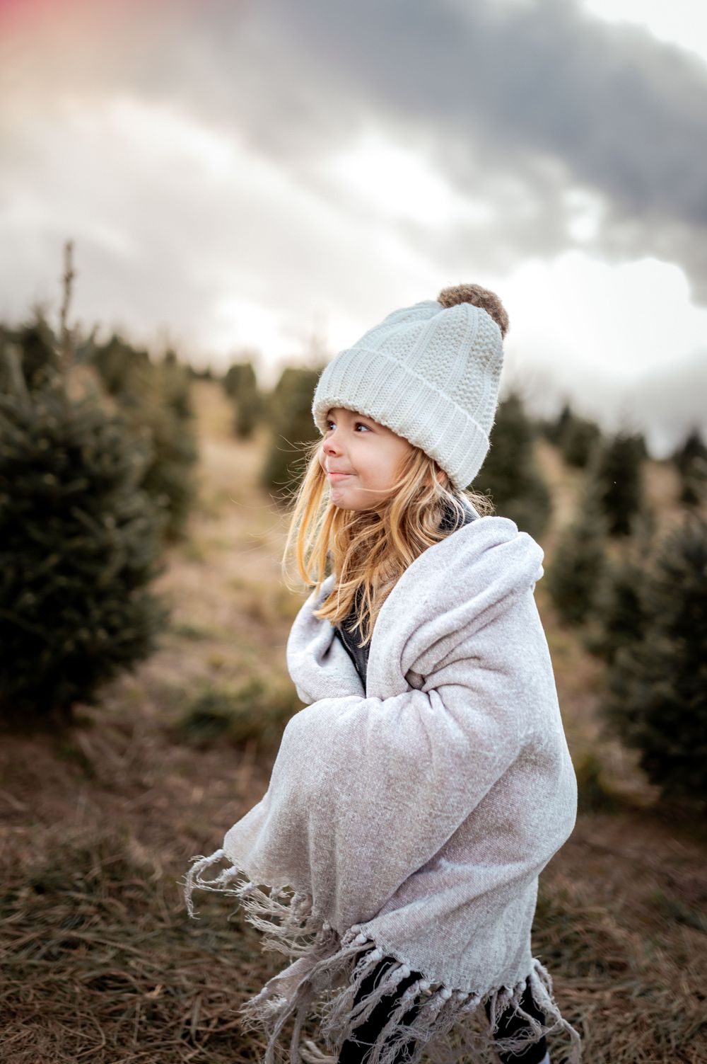Person wearing a white knit beanie and cozy blanket wrap poses thoughtfully among evergreen trees on a cloudy day.