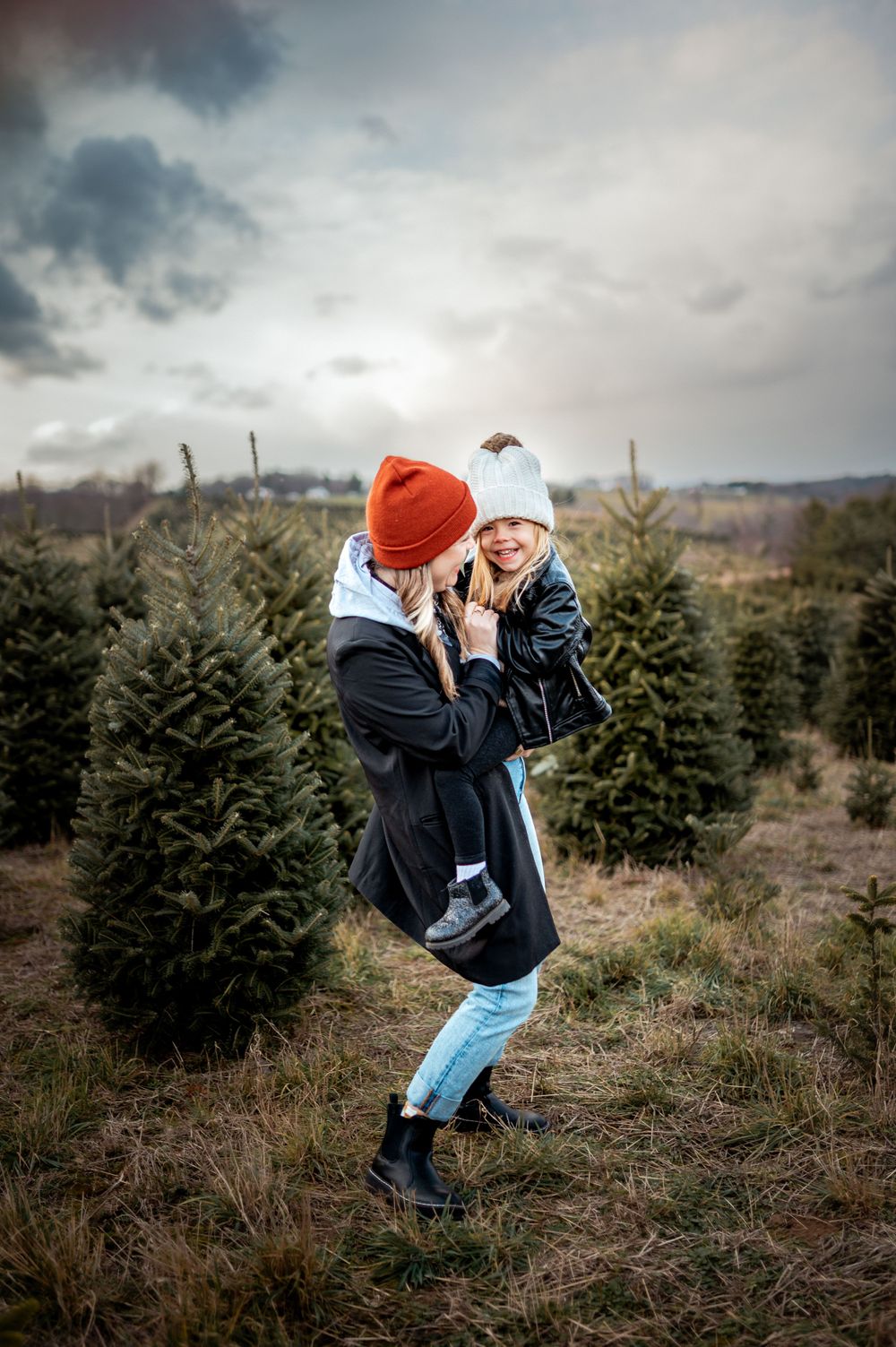 A parent wearing a red beanie and black jacket lifts a small child while standing among evergreen Christmas trees.