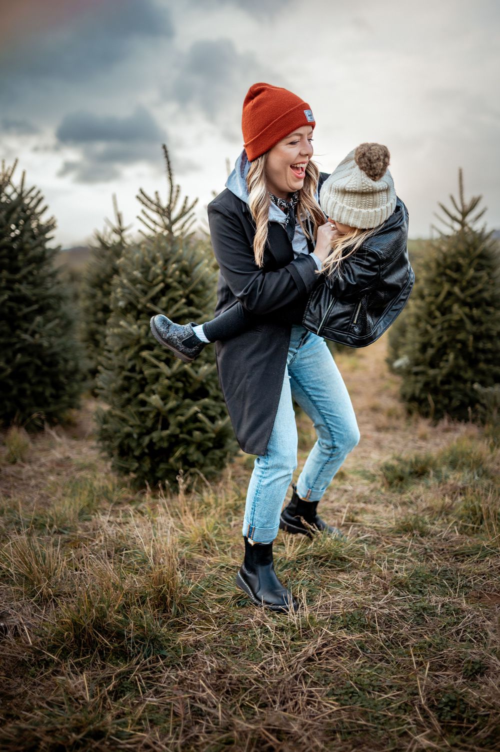 A couple in winter attire laughing together at a Christmas tree farm with evergreens in the background during cloudy weather.