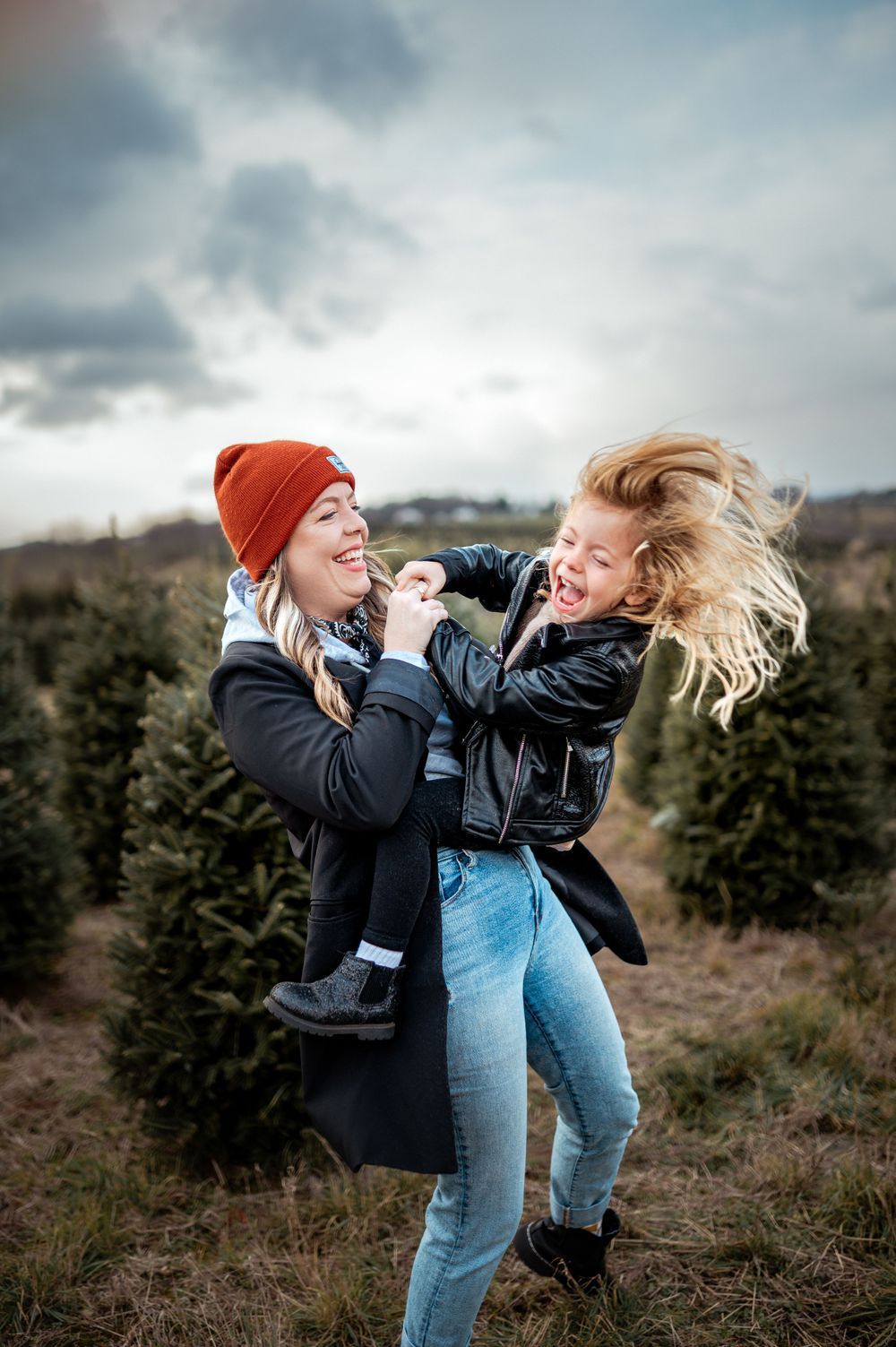 Two friends playfully interacting and laughing together at an outdoor Christmas tree farm on a cloudy winter day.