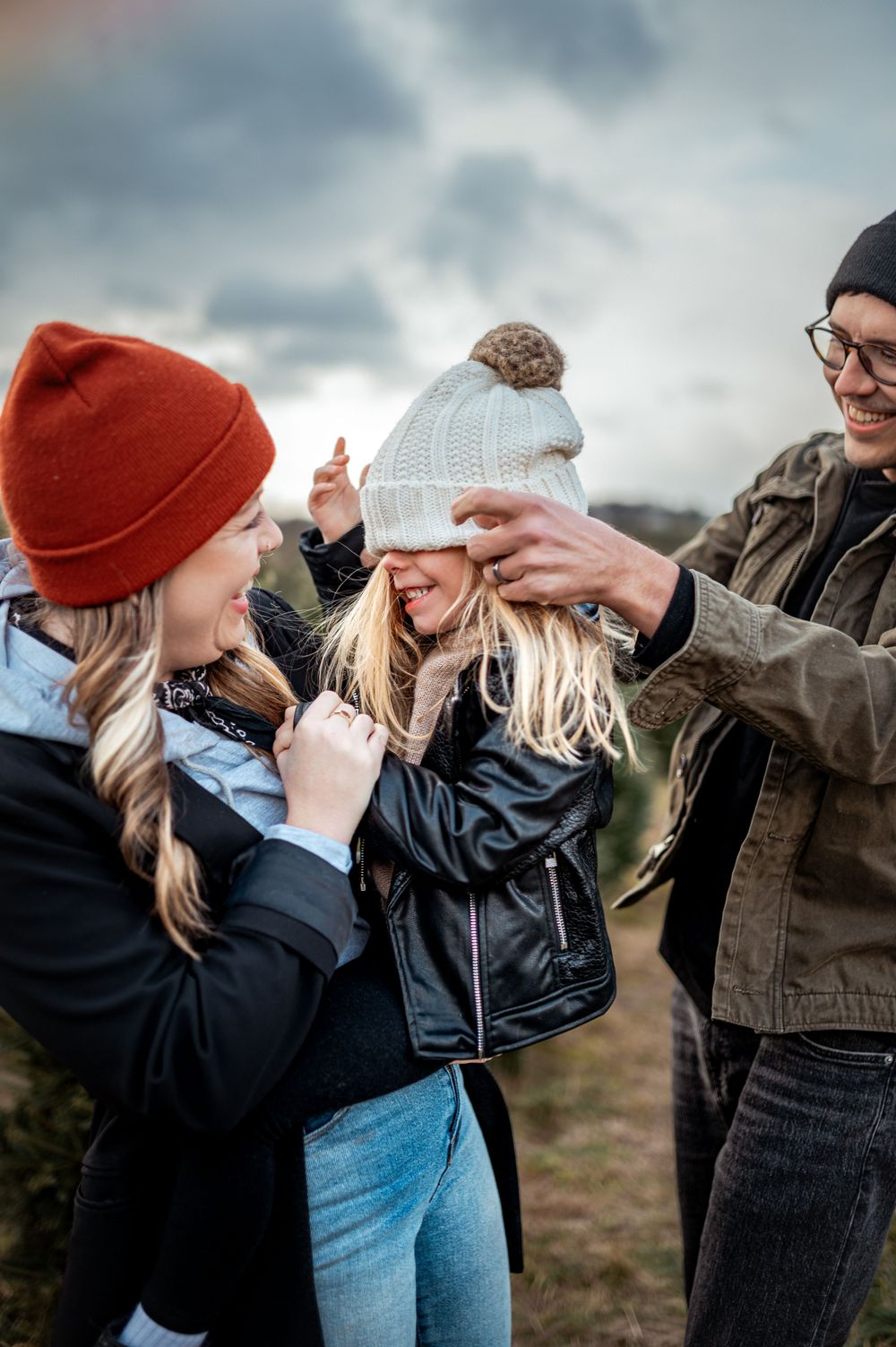 A playful moment captured outdoors as friends adjust a white knit winter hat against a moody cloudy sky.