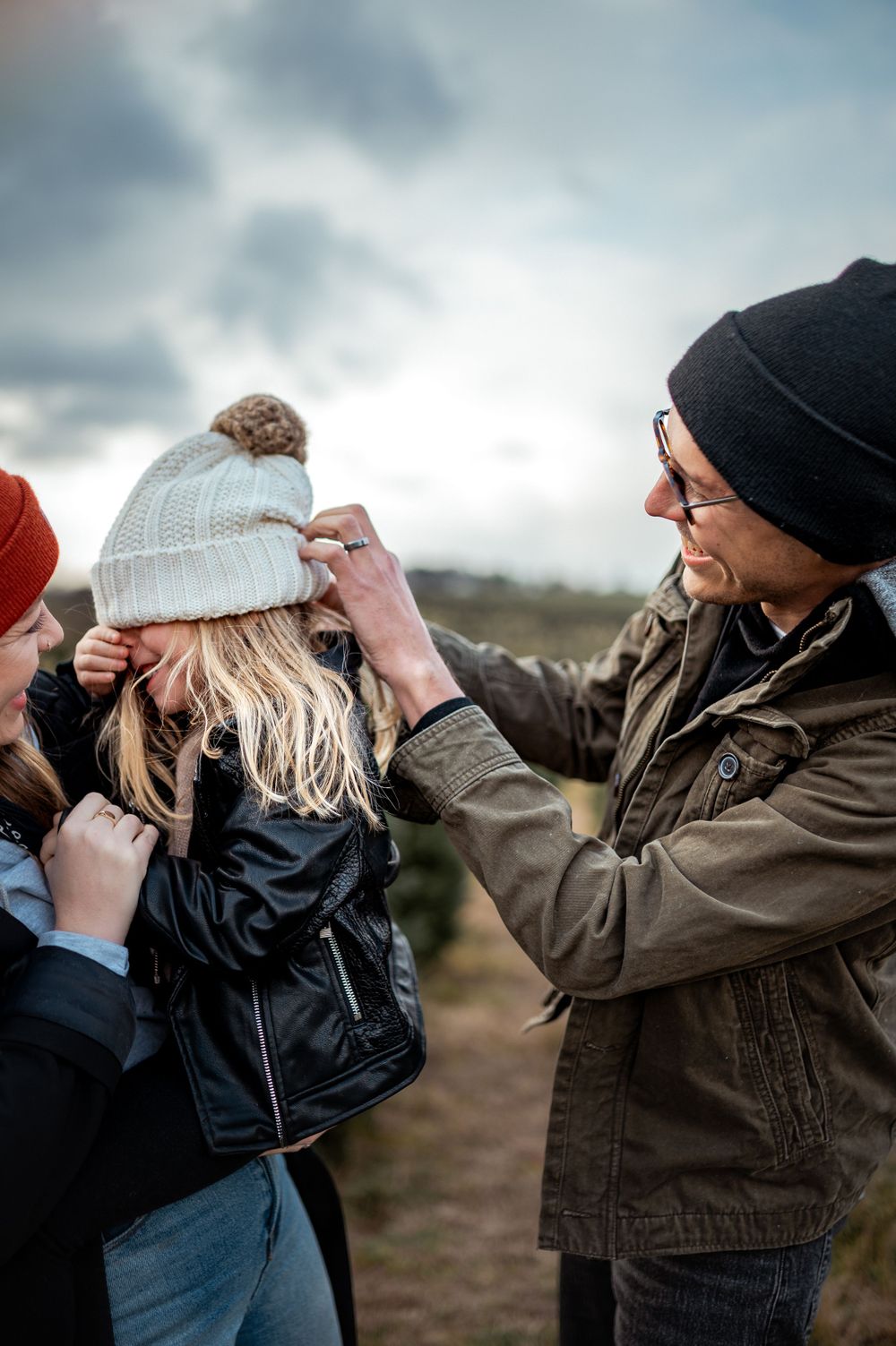 Three people sharing a lighthearted moment while adjusting a white pom-pom beanie outdoors on a cloudy day.