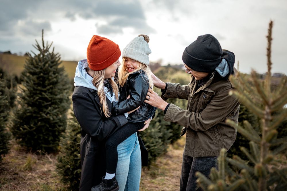 Three people in winter clothing sharing a laugh together while shopping for Christmas trees at an outdoor tree farm.