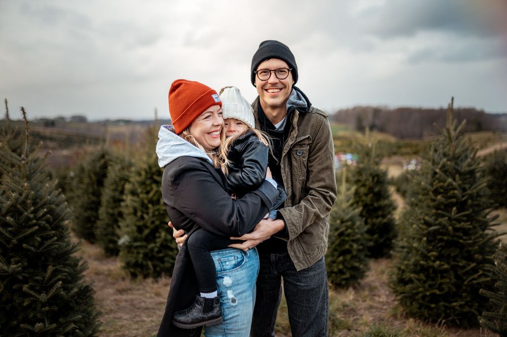 A happy couple shares a moment between rows of evergreen trees at a Christmas tree farm on an overcast day.