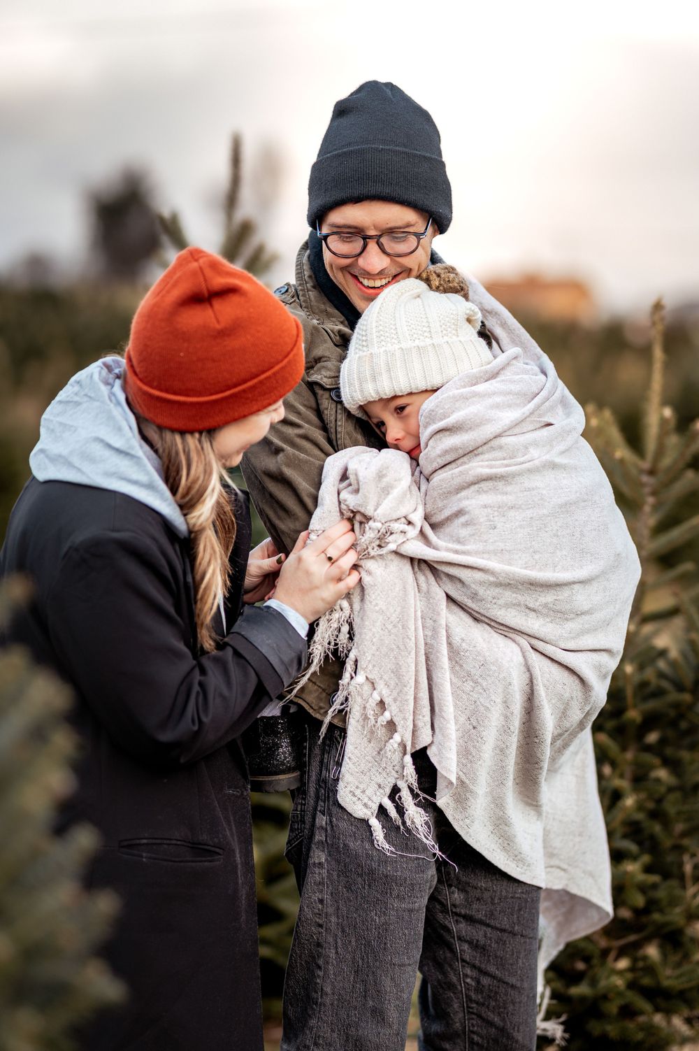A family shares a tender moment while wrapped in blankets at a Christmas tree farm on a cloudy day.
