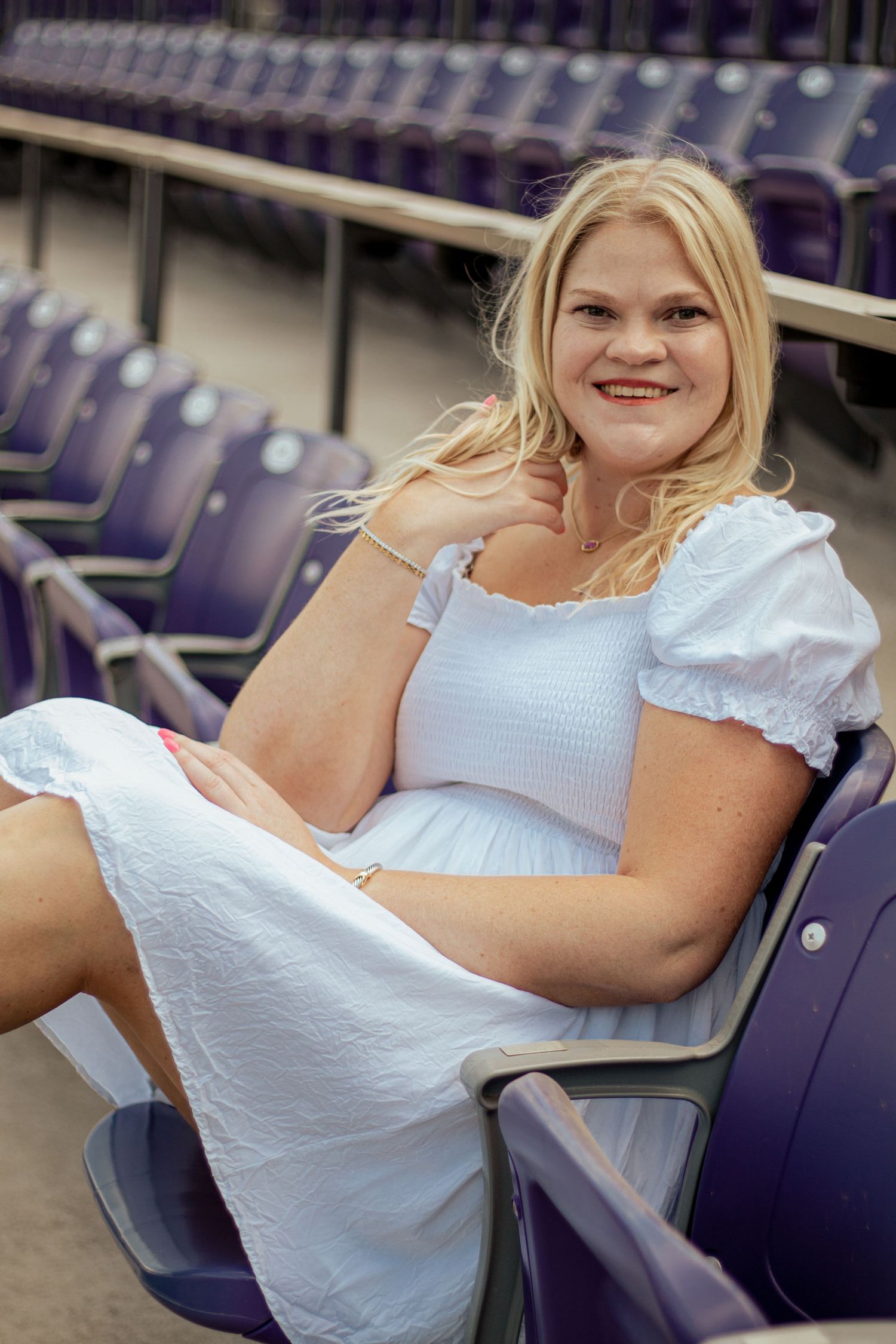 A woman in a white summer dress sits casually in a stadium seat surrounded by rows of purple chairs.