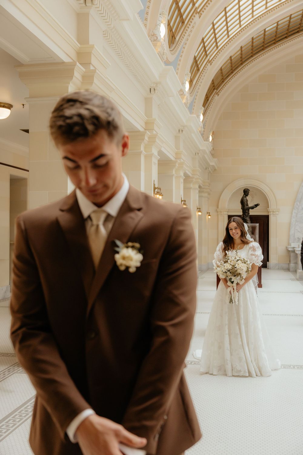 Groom in brown suit stands in foreground as bride in white gown approaches.