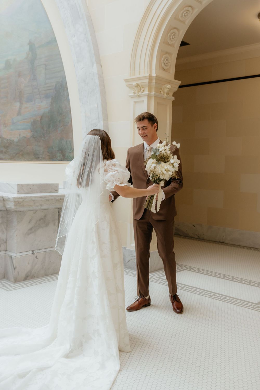Smiling groom in brown suit receives white floral bouquet from bride in lace wedding gown.