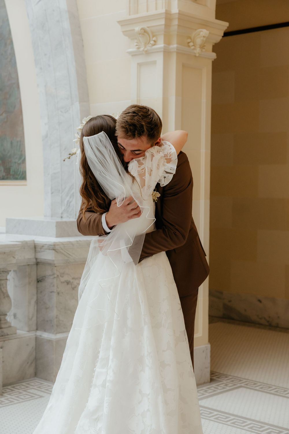 Newlyweds embrace tenderly in sunlit classical architecture hallway during wedding first look.