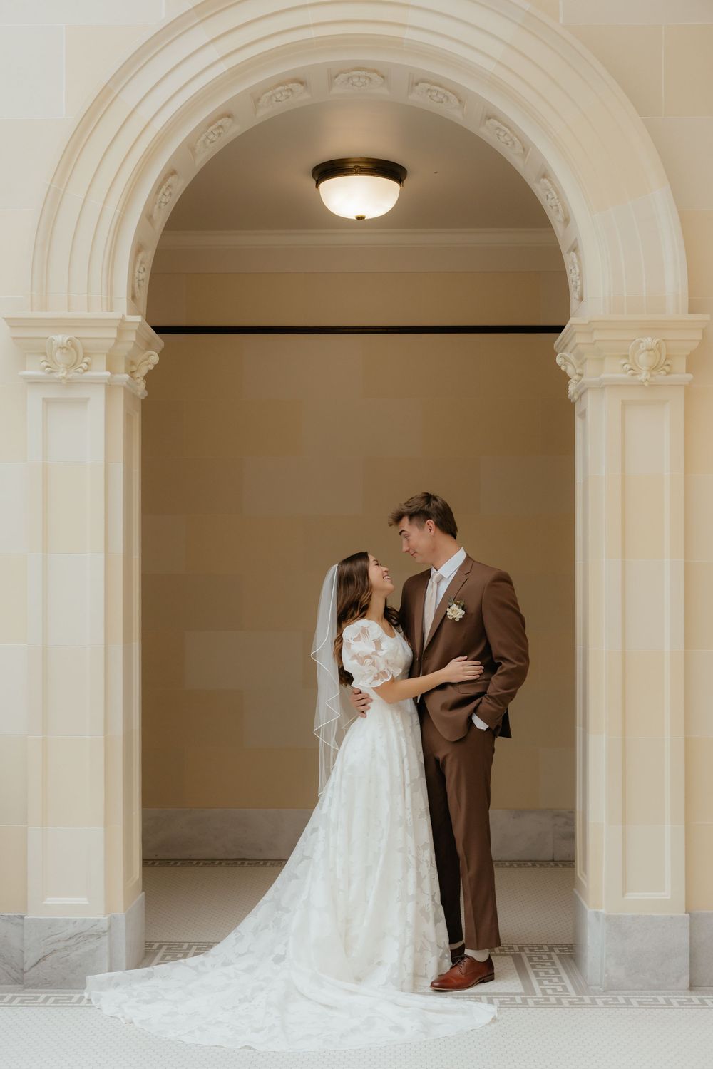 Wedding couple gazes lovingly at each other under ornate arch in golden-toned elegant venue.