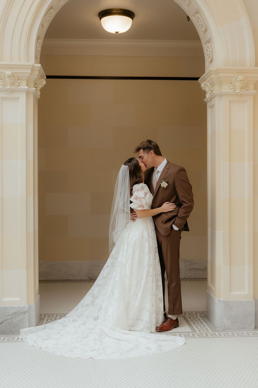 Groom kisses bride under beautiful arched doorway as her lace gown train flows on marble floor.