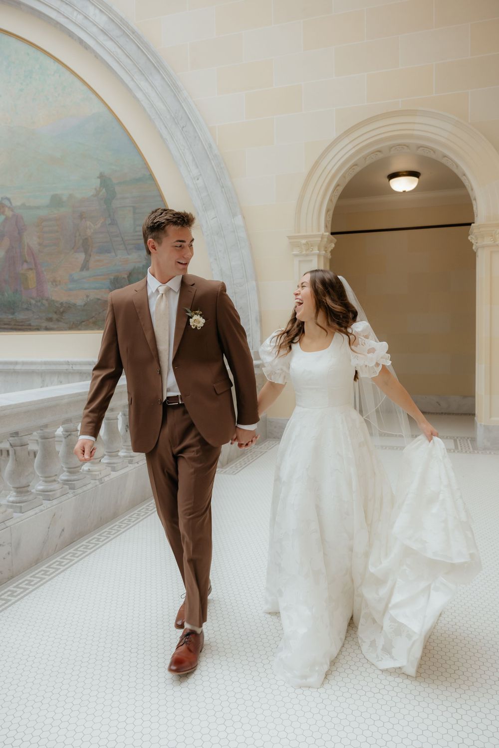 Smiling newlyweds walk hand in hand past a mural in a grand marble building interior.