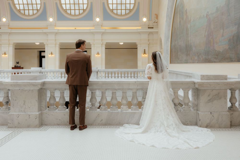 Bride and groom stand at ornate marble balcony overlooking grand hall in a historic building.