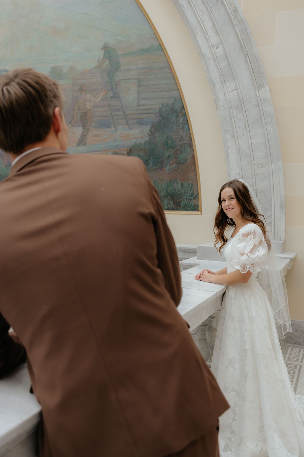 Bride in lace wedding dress smiles at groom in brown suit before a large mural in ornate venue.