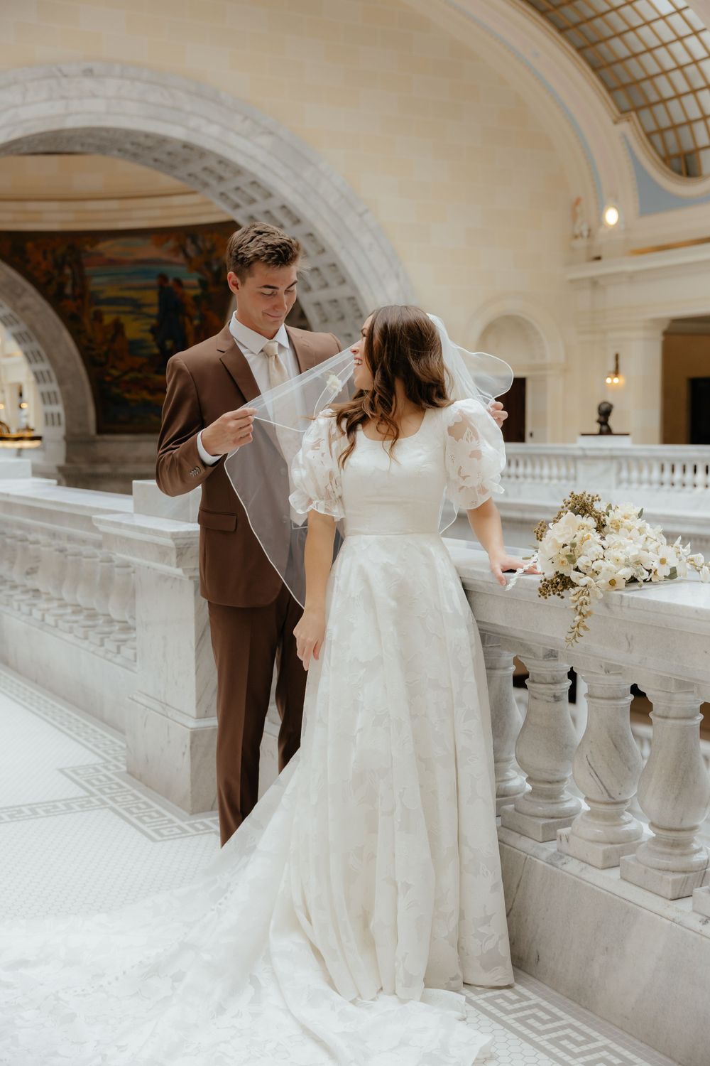 Groom in brown suit lifts bride's veil as she holds floral bouquet in grand marble building.