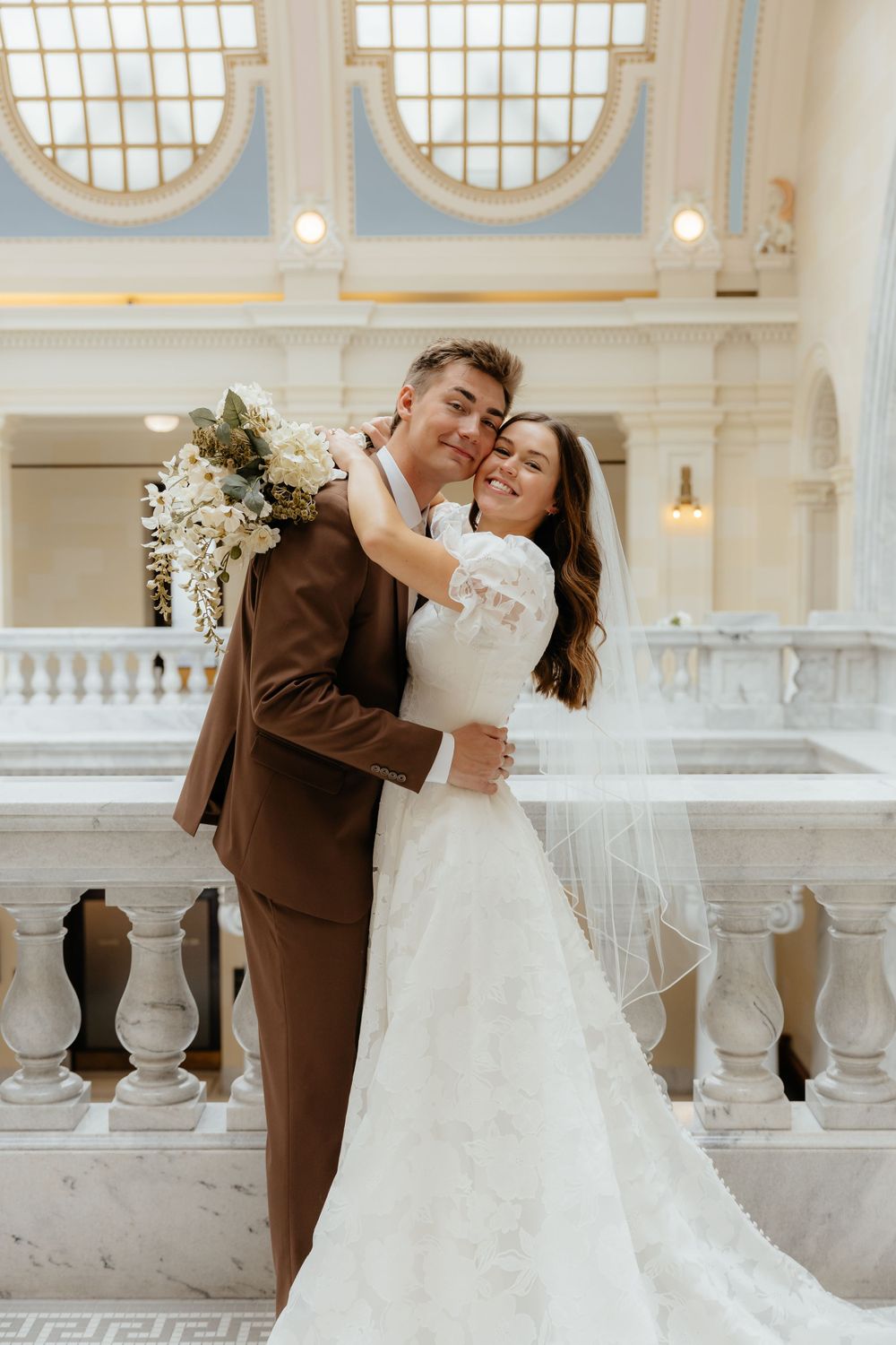 Joyful bride and groom embrace holding white floral bouquet inside grand marble wedding venue.