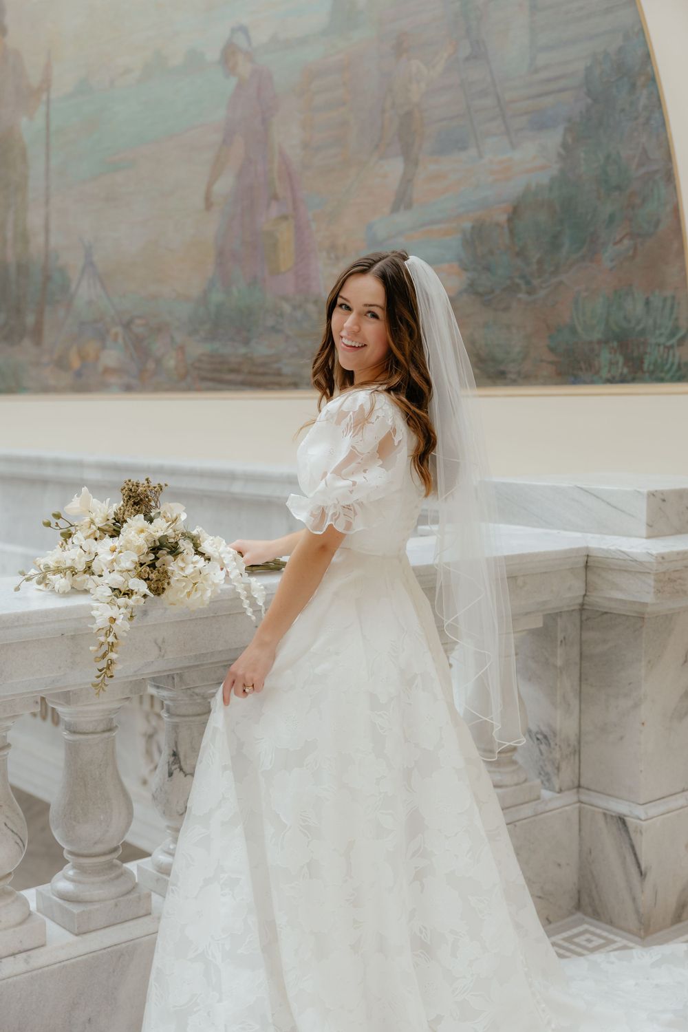 Smiling bride in white floral wedding dress and veil holds white bouquet near marble staircase.