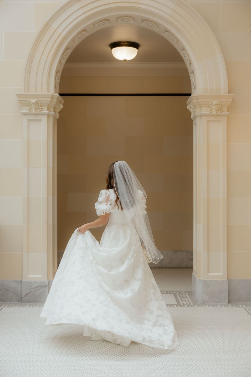 Bride in white wedding gown with veil walks through ornate arched doorway from behind.