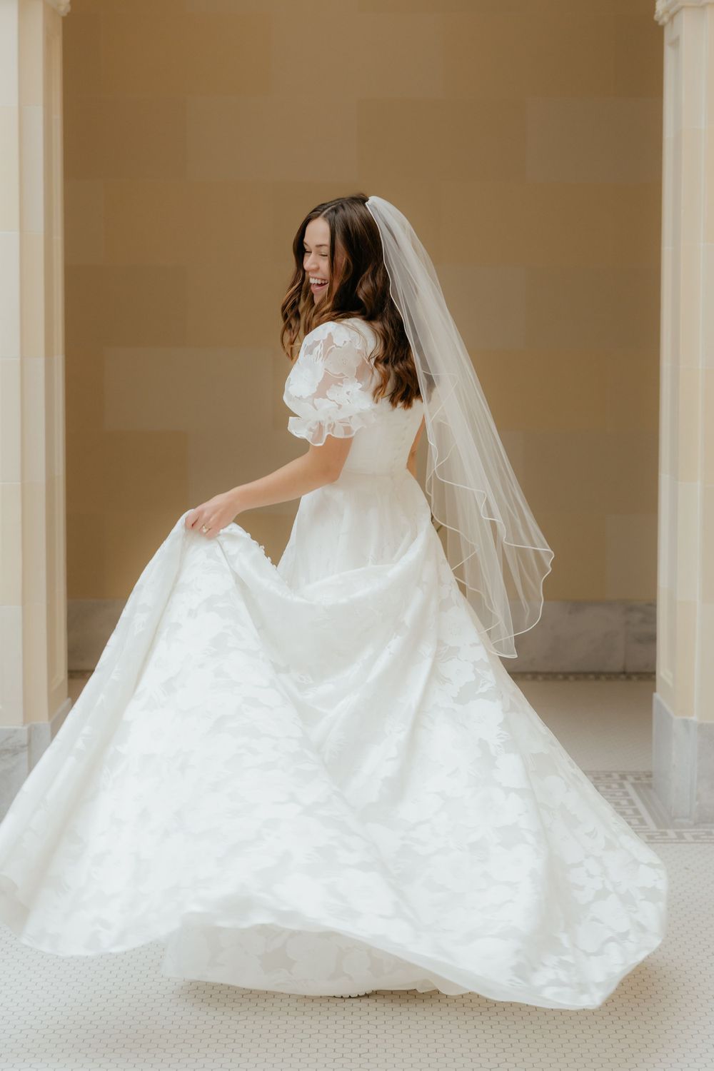 Bride in white floral wedding gown twirls skirt beside classical stone columns with veil.