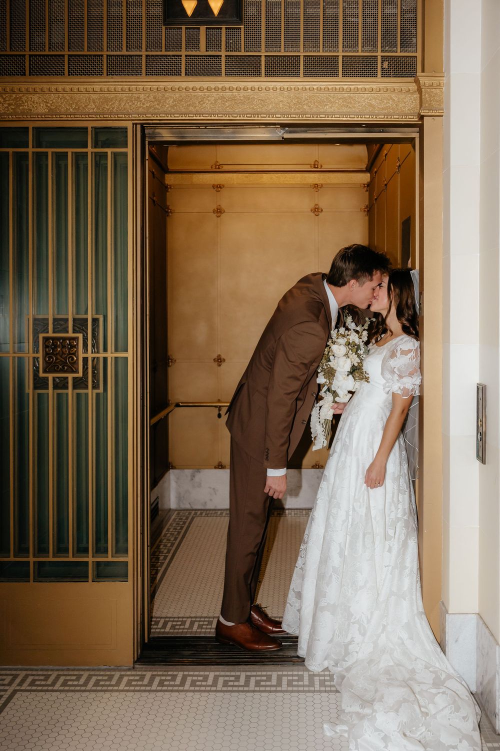 Bride and groom kiss in an ornate elevator doorway, bride holding white bouquet in lace gown.
