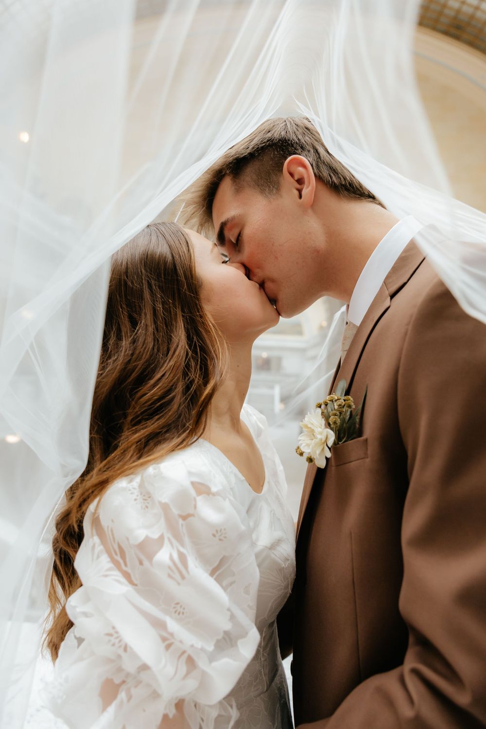 Bride and groom sharing a passionate kiss draped under a flowing white wedding veil.