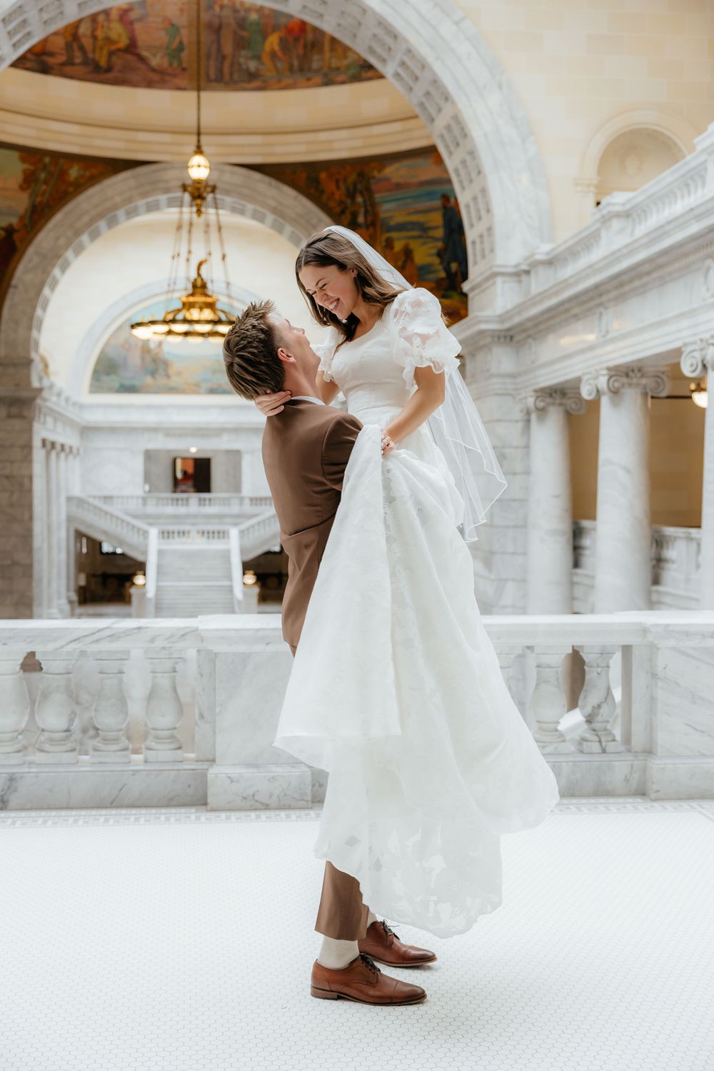 Groom lifting joyful bride in white wedding dress inside stunning marble domed capitol building.