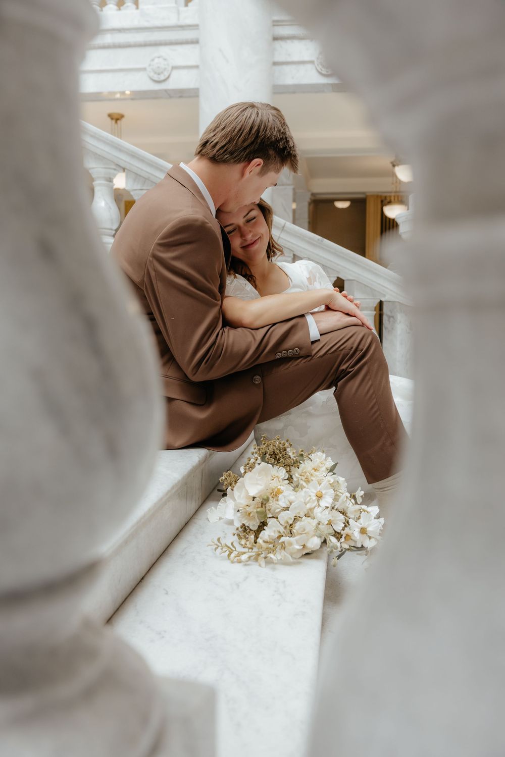 Bride and groom sharing a romantic embrace on marble stairs with elegant white floral bouquet.