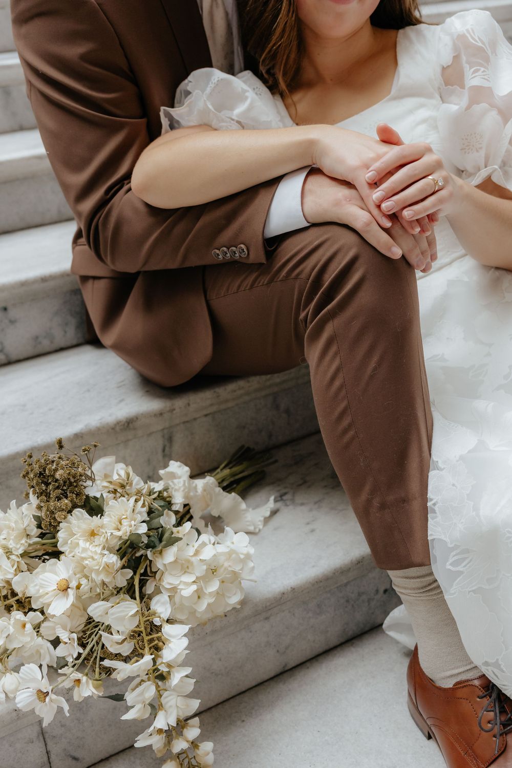 Close-up of bride and groom holding hands on marble steps with white floral bouquet beside them.