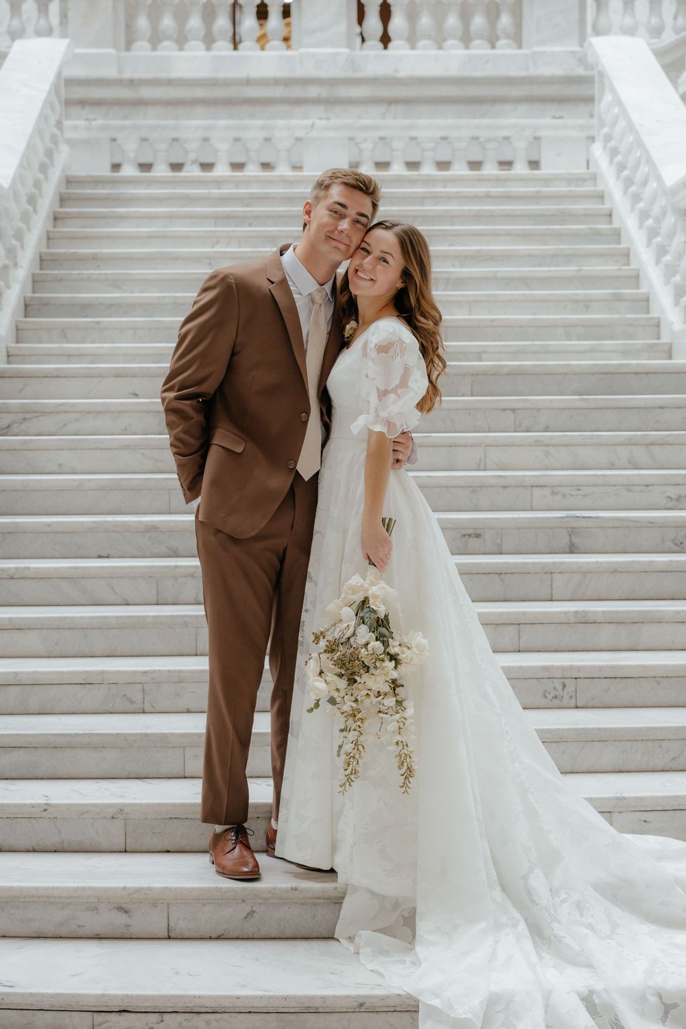 Bride and groom posing together on marble staircase, bride holding cascading white floral bouquet.
