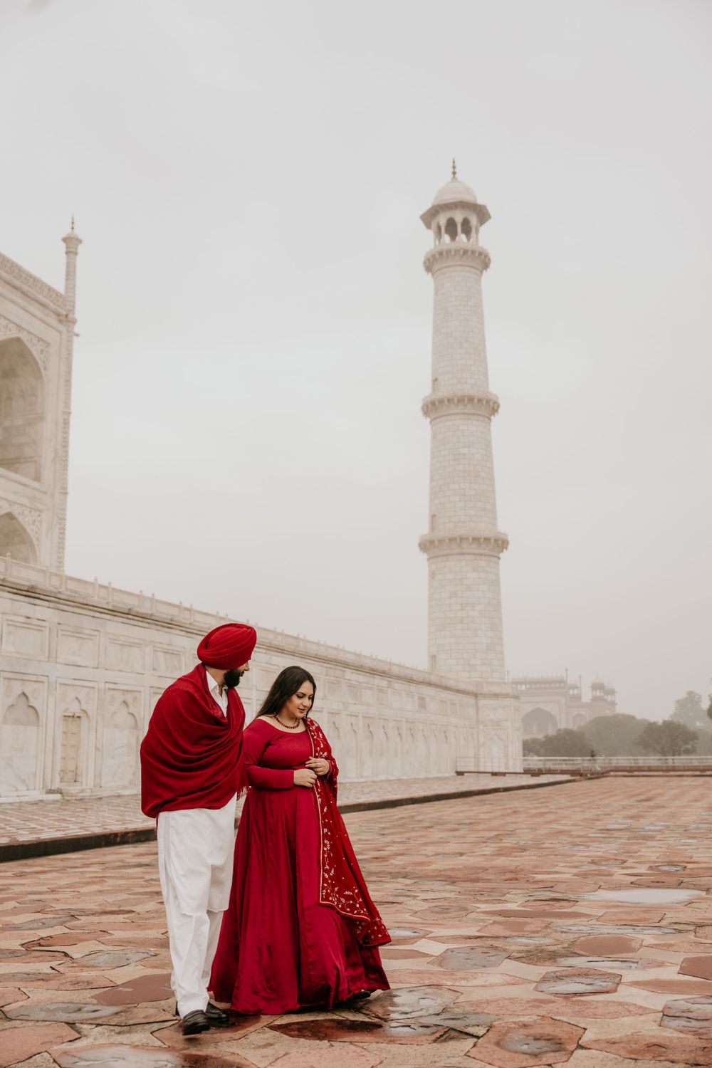 A couple in matching red outfits stands together at the iconic Taj Mahal on a misty morning.