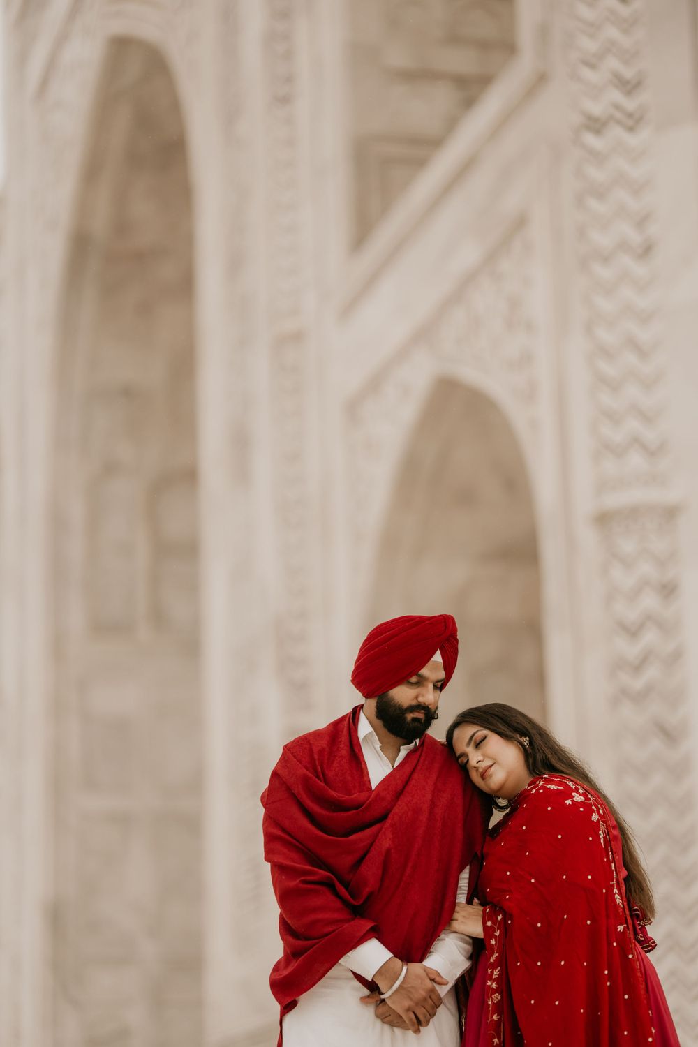 A couple in traditional red Indian attire share an intimate moment against an ornate white architectural backdrop.