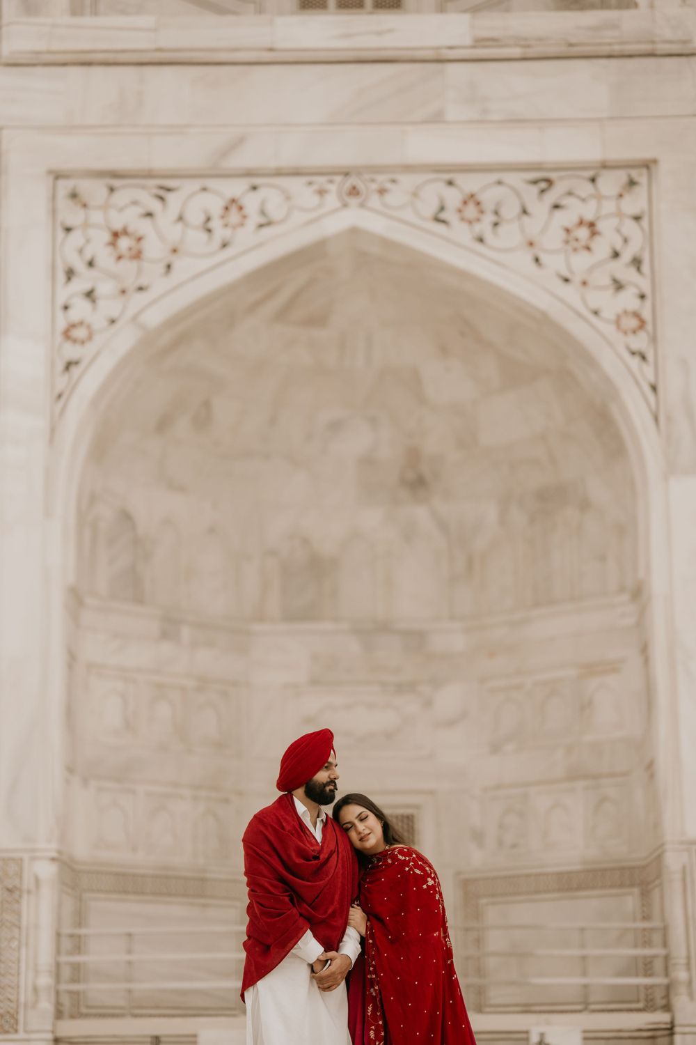 A couple in matching red traditional Indian attire embraces under an ornate white marble archway at a historic monument.