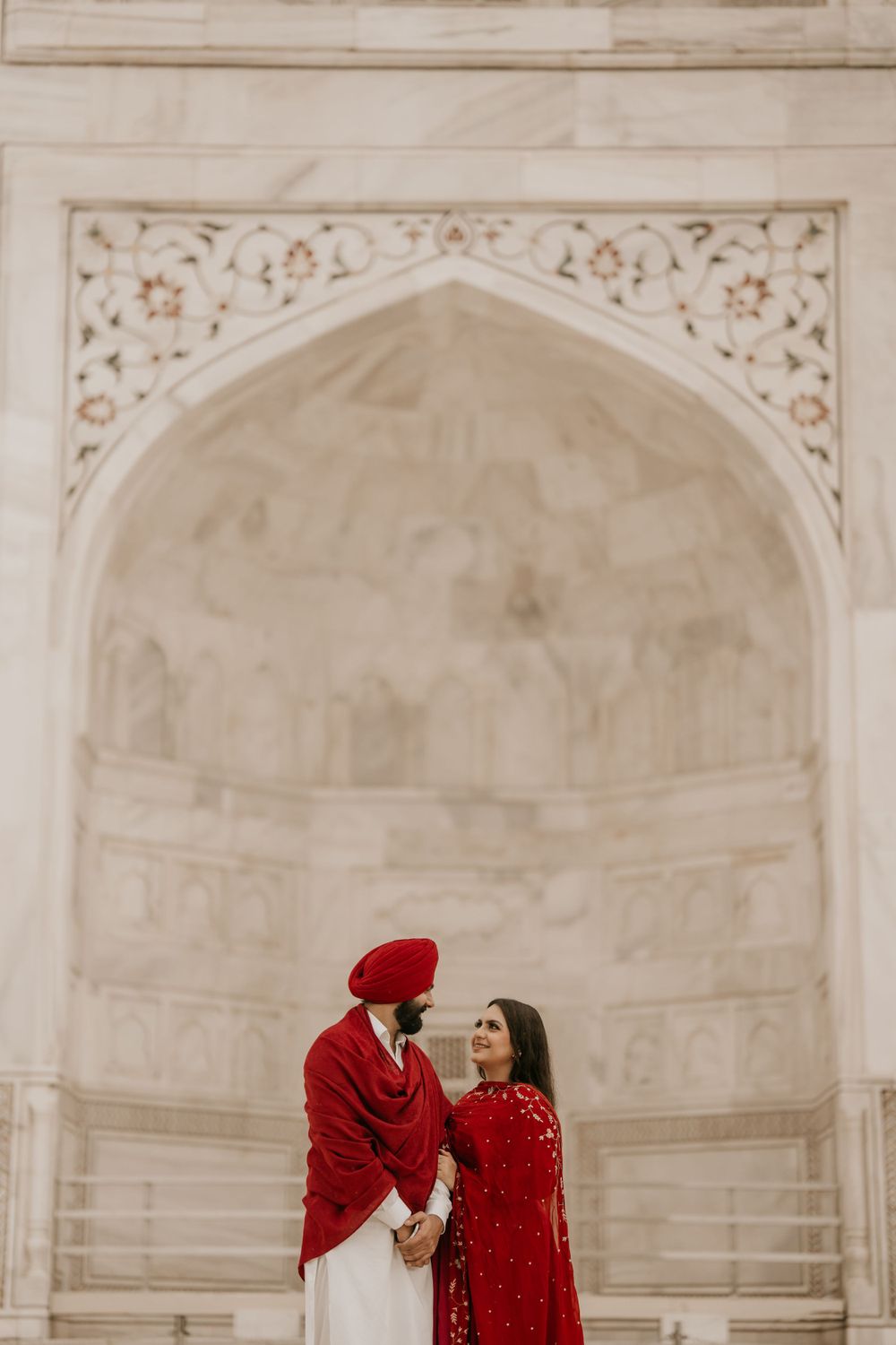 A couple in traditional red Indian attire holds hands while standing in front of an ornate marble archway.