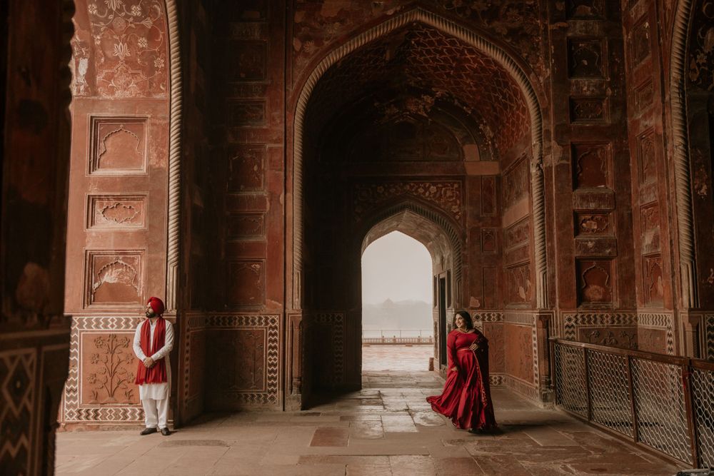A person in a red dress stands beneath ornate archways of an ancient Indian palace with intricate stone carvings.