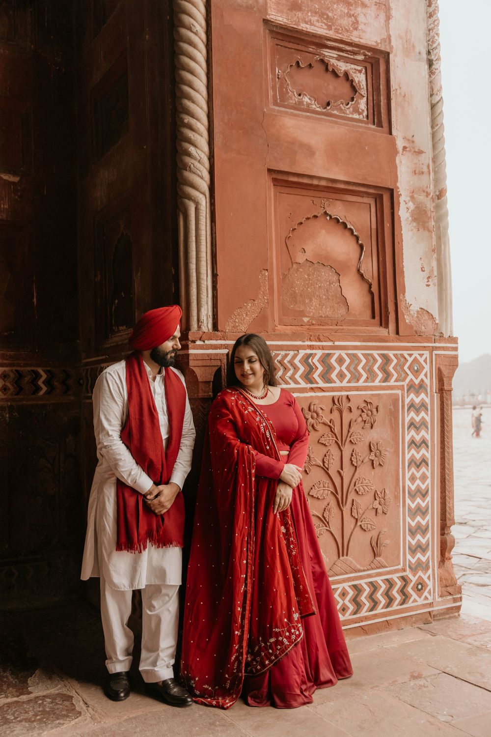 A couple wearing traditional red and white Indian wedding attire poses against an ornate terracotta wall with decorative patterns.