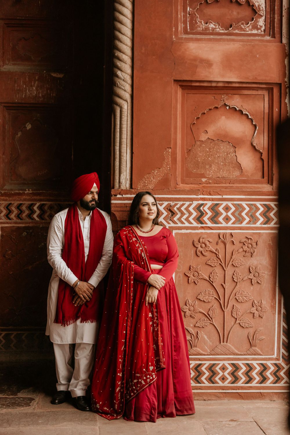Traditional Indian wedding couple in red and white attire posing against ornate terracotta wall with carved details and patterns.