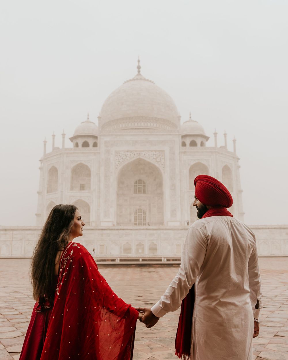 A couple in traditional Indian attire holding hands near the misty Taj Mahal in Agra.