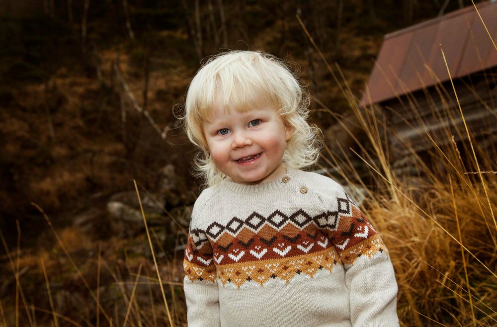 Blond 2-åring med strikkegenser ser i kamera og ler på utendørsfotografering i Farsund