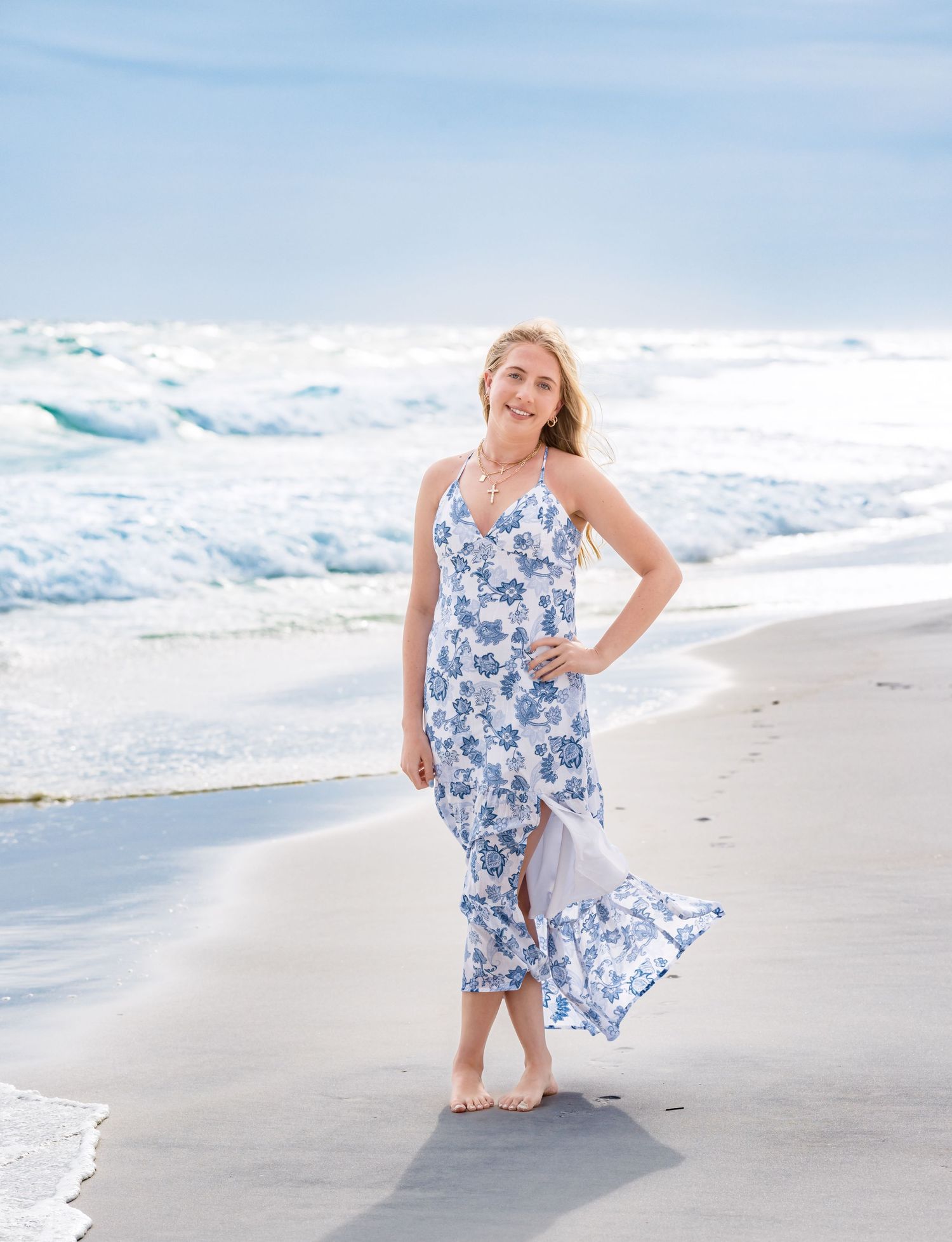 A person in a blue and white floral sundress poses on a sunny beach with waves crashing in the background.