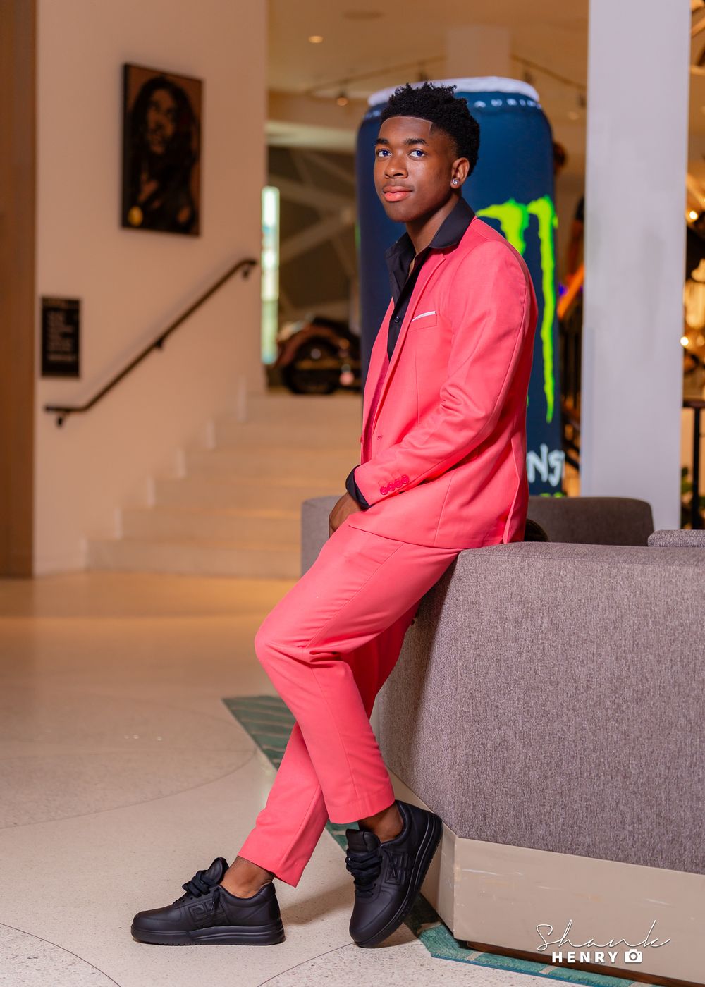 A person poses on a grey couch in a modern interior space wearing a vibrant coral-colored suit and black dress shoes.