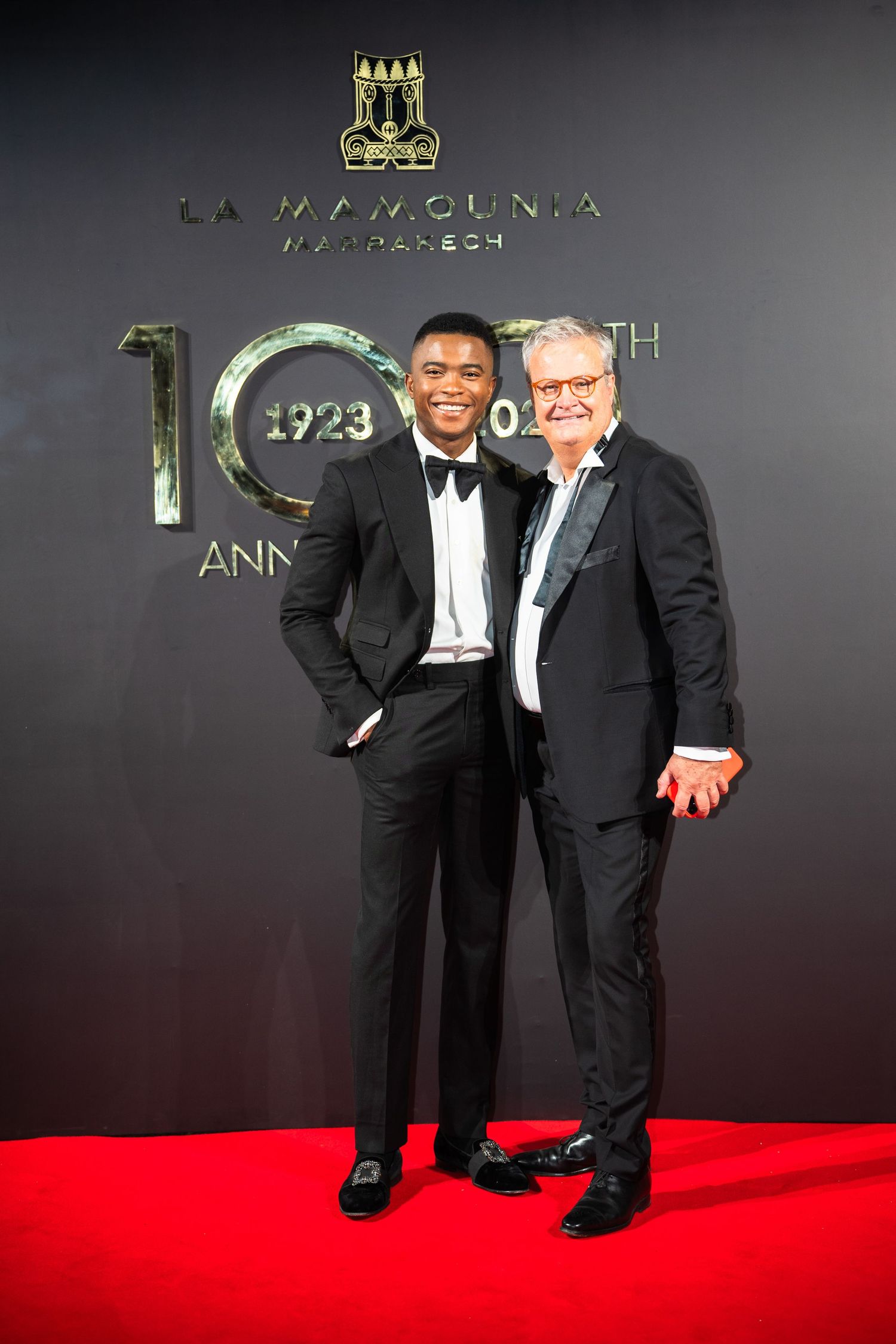 Two guests in formal black tuxedos pose together on a red carpet at a 100th anniversary celebration event.