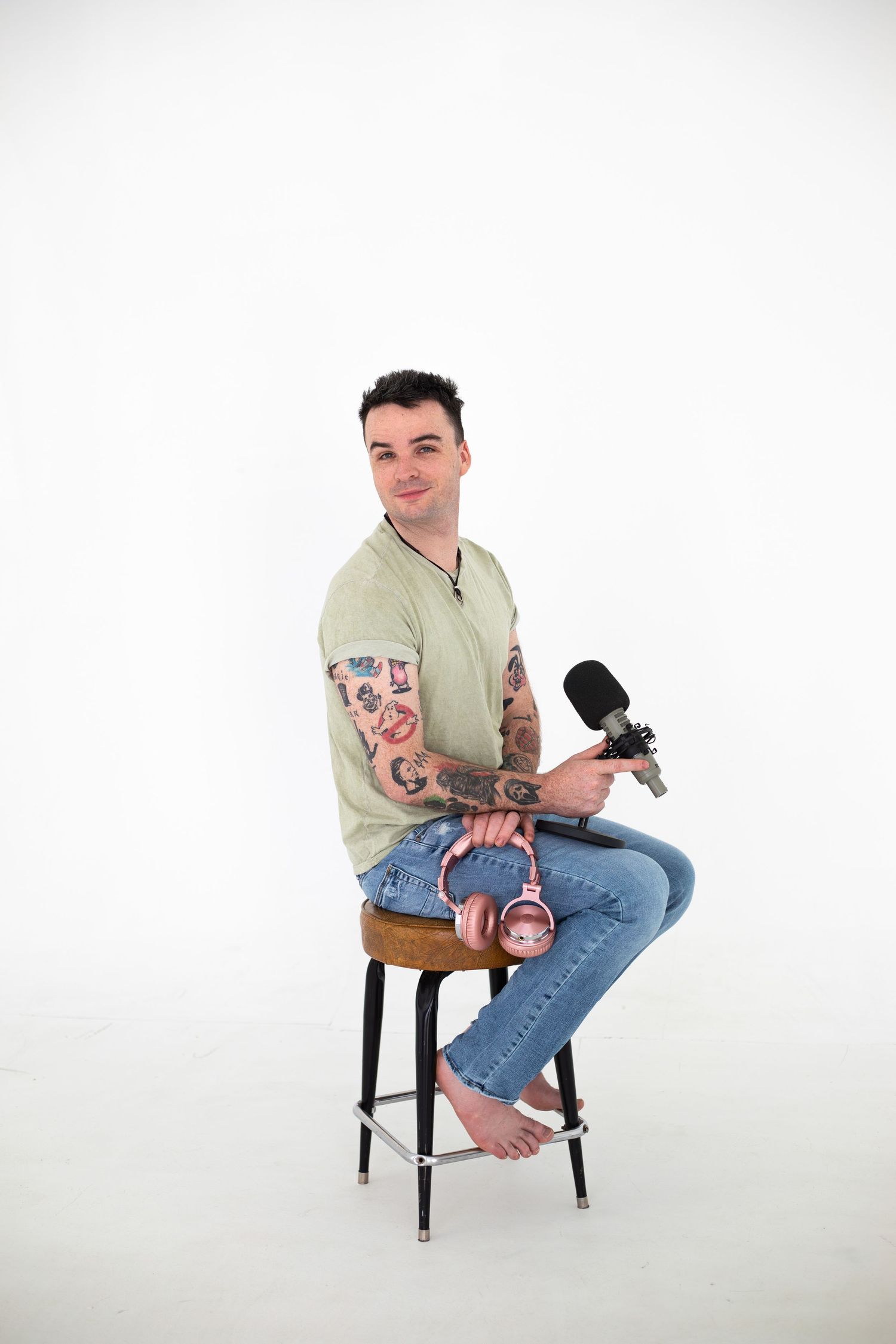 Professional podcast host with tattoos sits on bar stool holding microphone in studio setting.