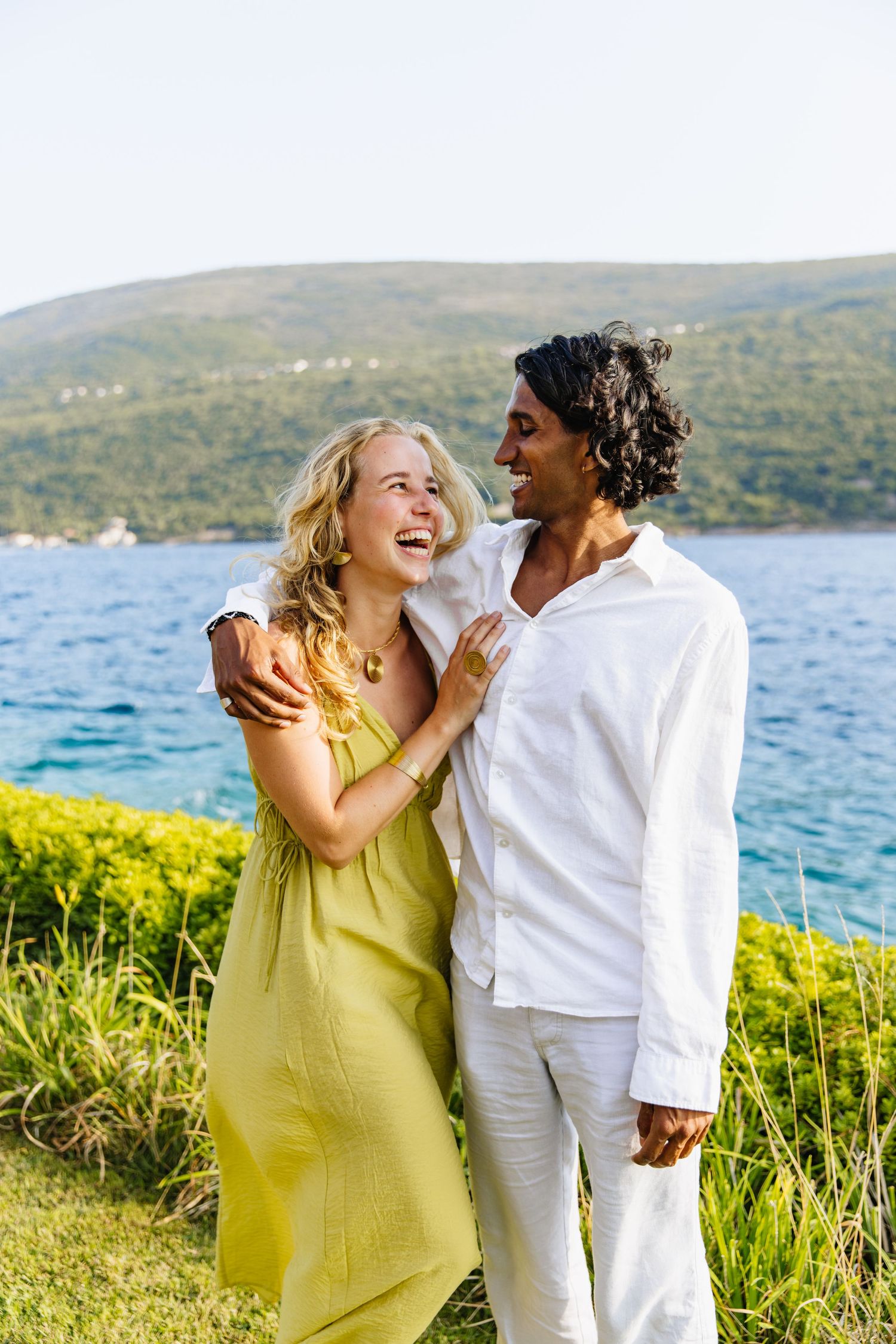 Couple in summer attire sharing a joyful moment together by the seaside with blue water and hills in background.
