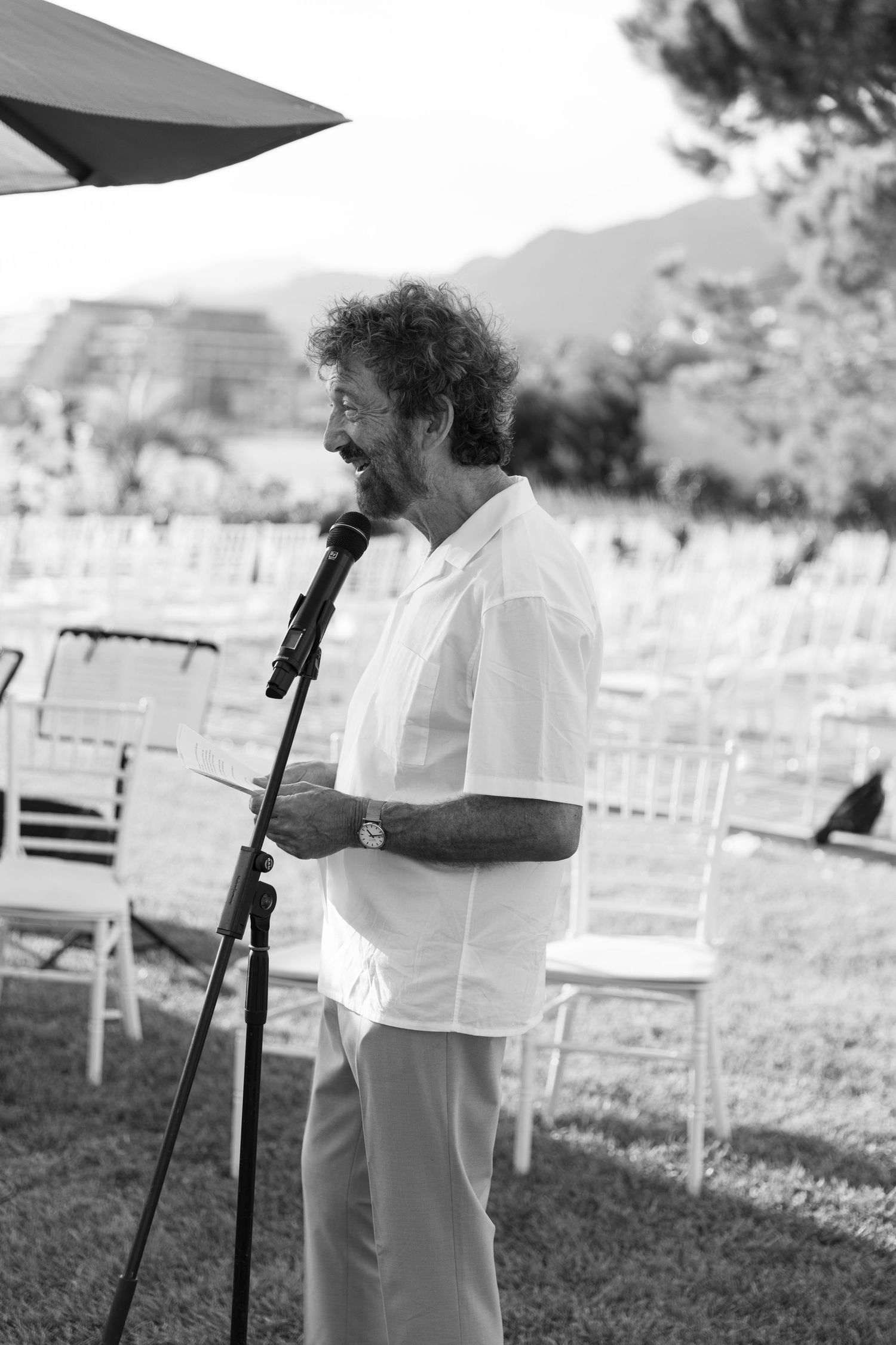 Someone speaks into a microphone at an outdoor event with white chairs and a garden setting in black and white photography.