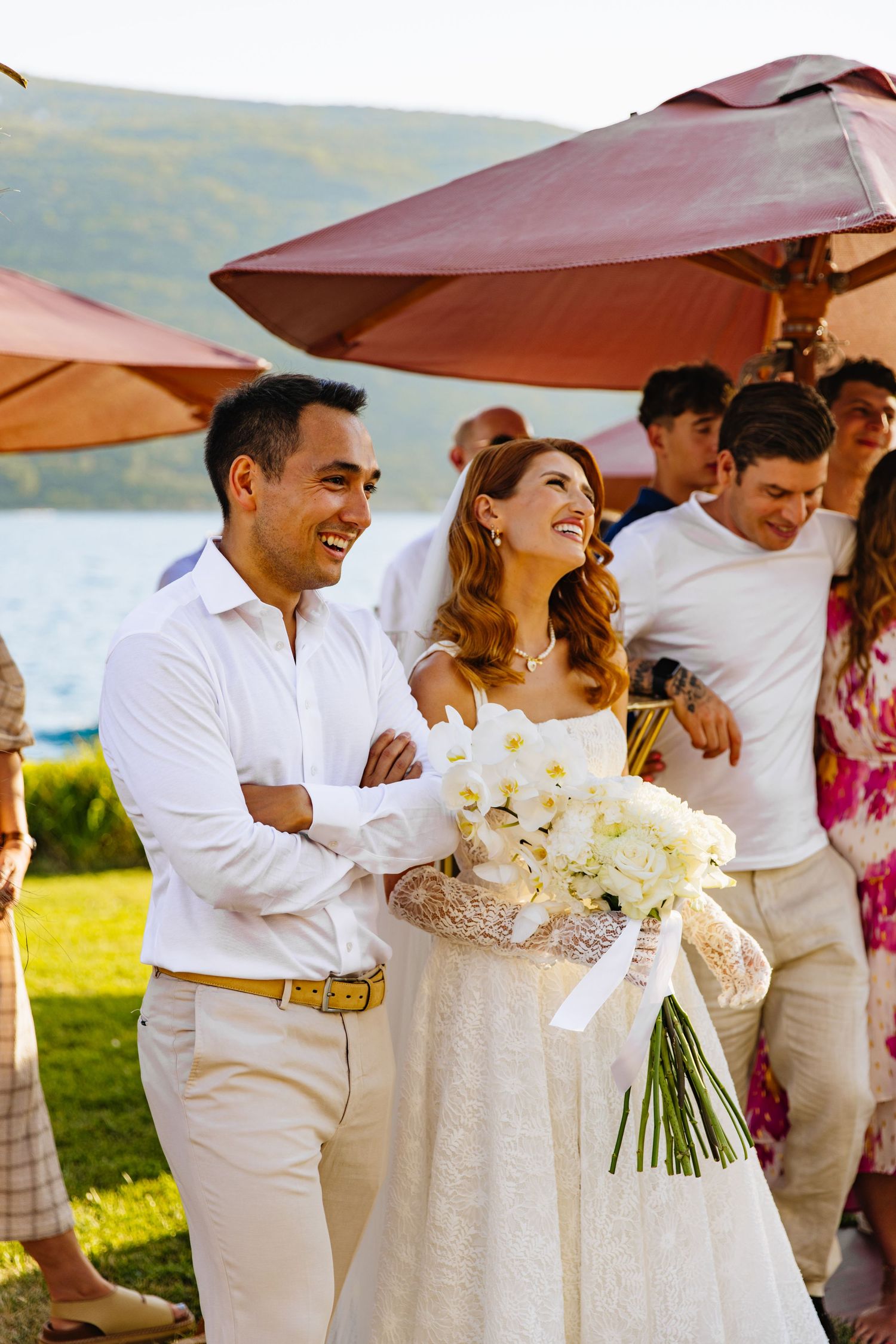 A wedding celebration on a scenic lakeside lawn with guests in white and floral attire beneath orange umbrellas.