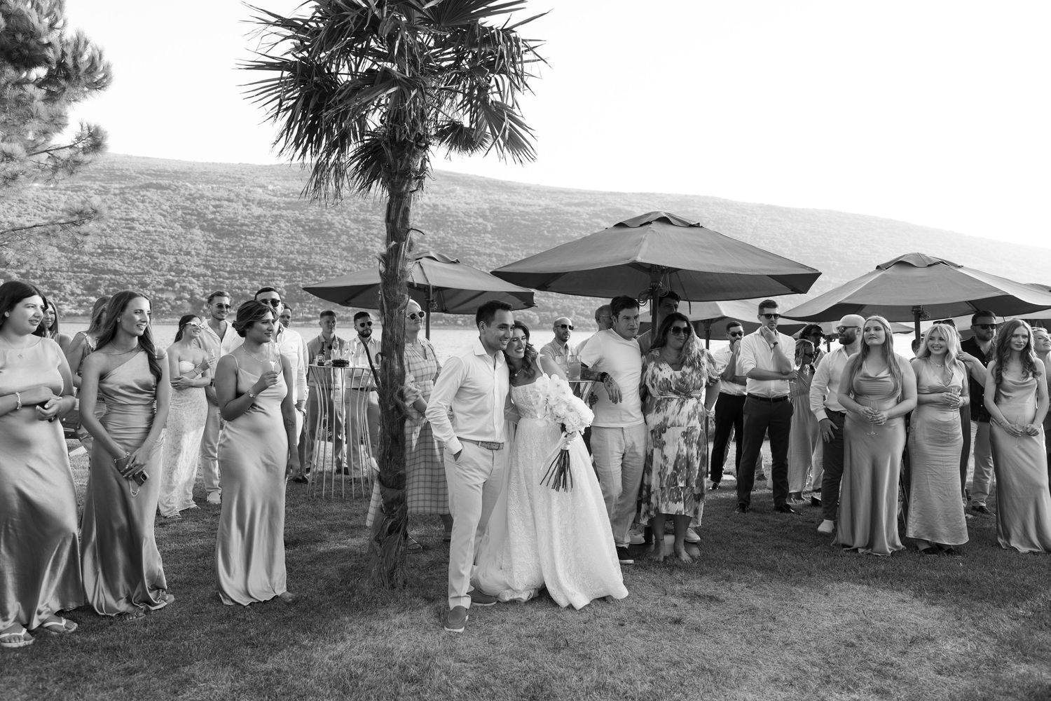 A black and white photograph of wedding guests gathered outdoors under umbrellas and palm trees on a hillside.